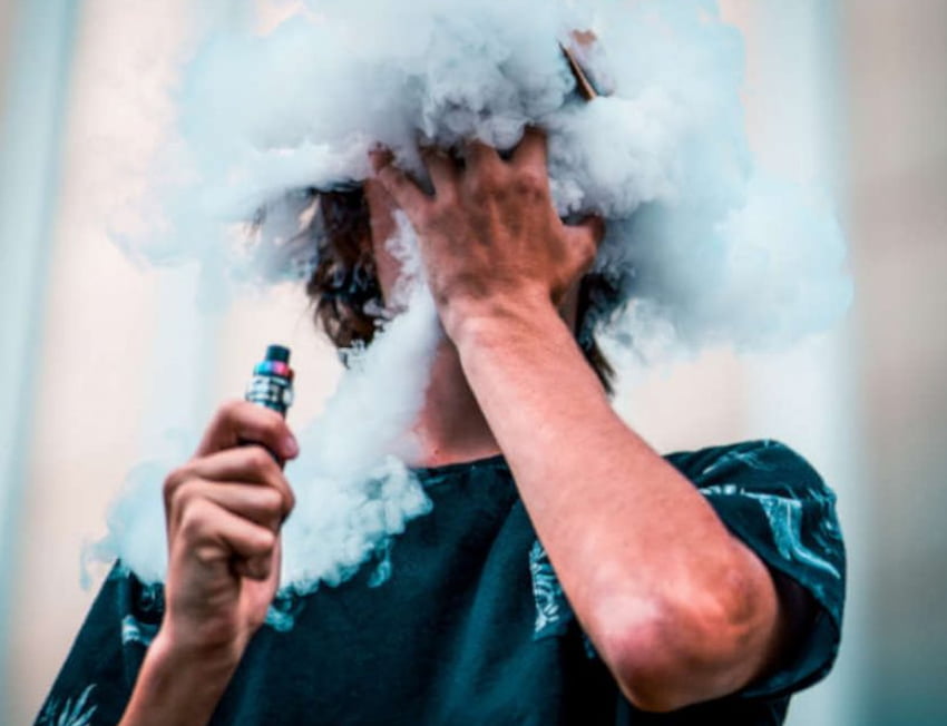 homem com um vape na mão esquerda tampando o rosto com a mão direita