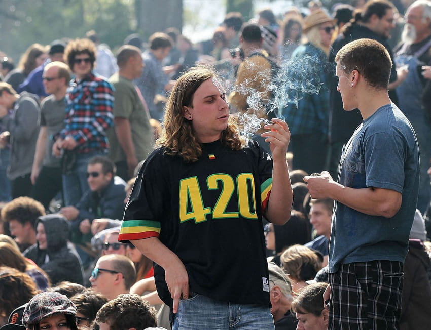 imagem com várias pessoas. Em foco um homem fuma maconha com uma camiseta escrita 420